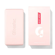 Glossier Body Hero Exfoliating Bar 141 g - jasnoróżowa kostka z wytłoczonym napisem Glossier obok różowego pudełka z napisem Body Hero, białe tło | Ellemania