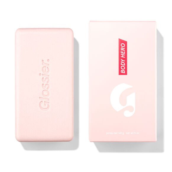 Glossier Body Hero Exfoliating Bar 141 g - jasnoróżowa kostka z wytłoczonym napisem Glossier obok różowego pudełka z napisem Body Hero, białe tło | Ellemania