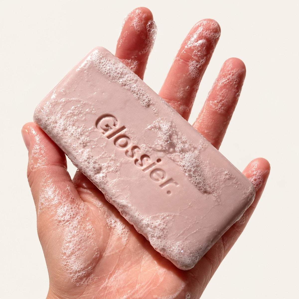 Glossier Body Hero Exfoliating Bar 141 g - kostka z wytłoczonym logo w otwartej dłoni, pokryta pianą, jasne tło | Ellemania