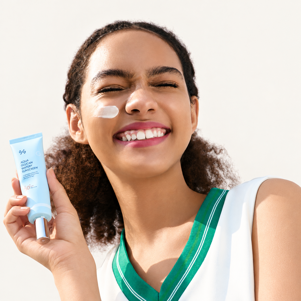 Aqua Glucan Watery Sunscreen - uśmiechnięta modelka z kremem na policzku trzyma niebieską tubkę z metalową końcówką | Ellemania