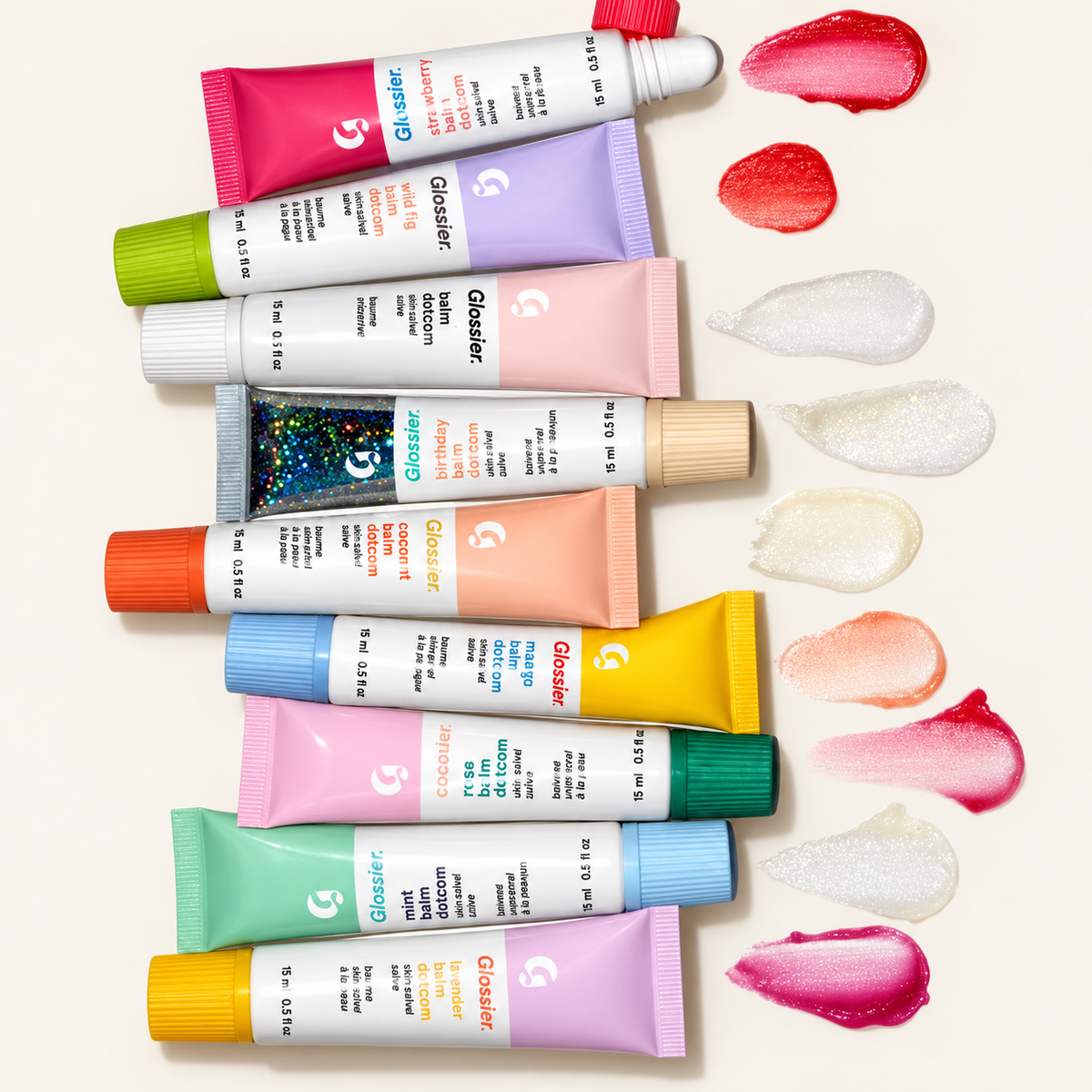 Glossier Balm Dotcom - dziewięć tubek 15 ml (m.in. strawberry, wild fig, birthday, mango, rose, mint, coconut, lavender) ułożonych obok smug produktu w różnych kolorach, na jasnym tle | Ellemania