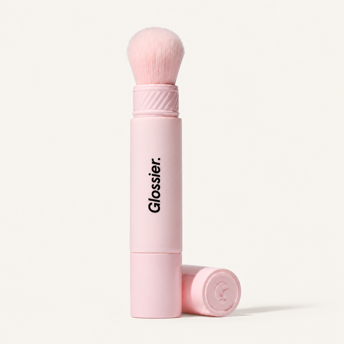 Glossier Cloud Paint Dual-ended Cheek Blush Brush - różowy dwustronny pędzel z miękkim włosiem i czarnym napisem Glossier na trzonku, obok leży nakrętka z logo, na jasnym tle | Ellemania