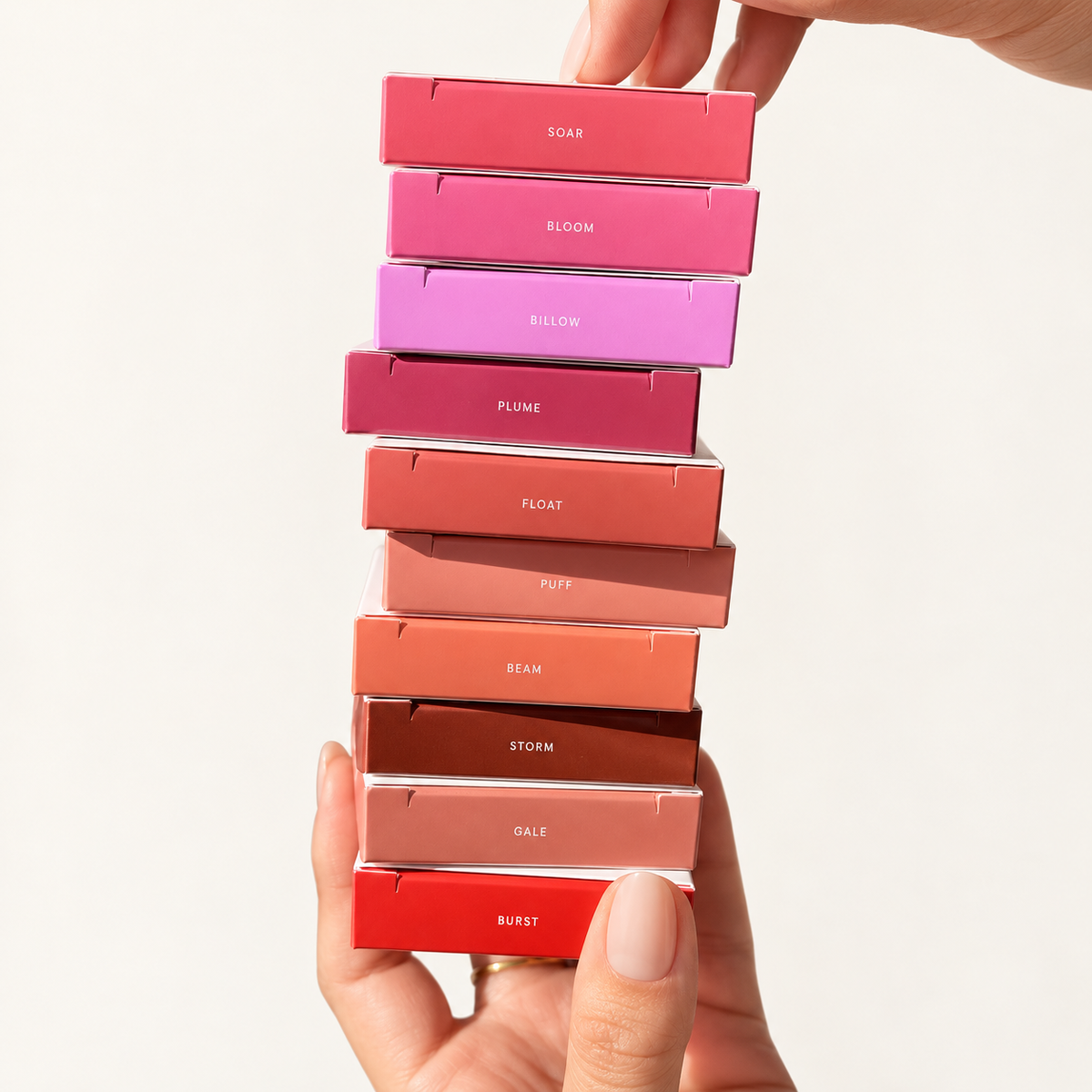 Glossier Cloud Paint Plush Blush - stos dziesięciu pudełek w różnych odcieniach z podpisami Soar, Bloom, Billow, Plume, Float, Puff, Beam, Storm, Gale i Burst, trzymany w dłoniach na jasnym tle | Ellemania
