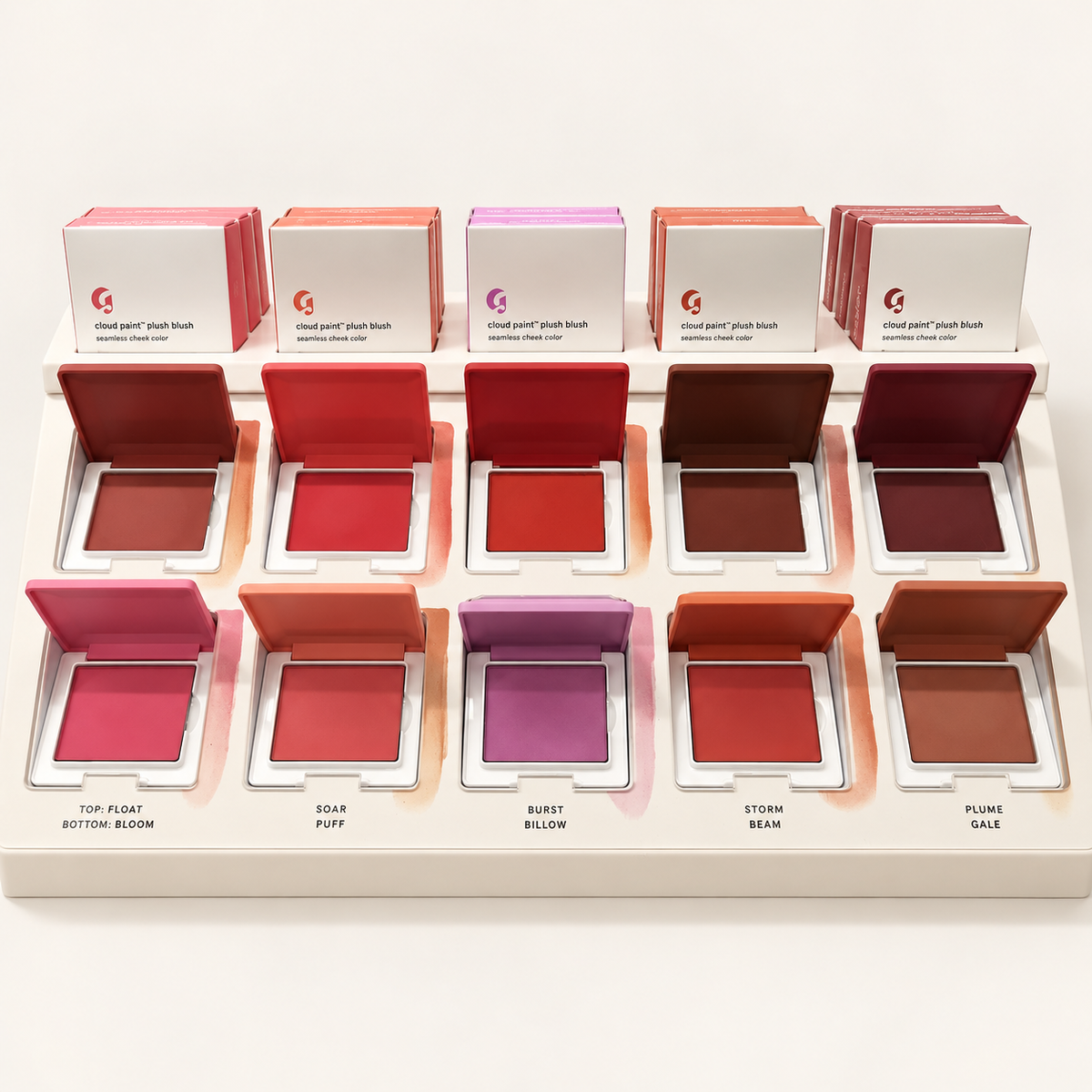 Glossier Cloud Paint Plush Blush - ekspozytor z dziesięcioma otwartymi kompaktami w różnych odcieniach i rzędem pudełek na jasnym tle | Ellemania