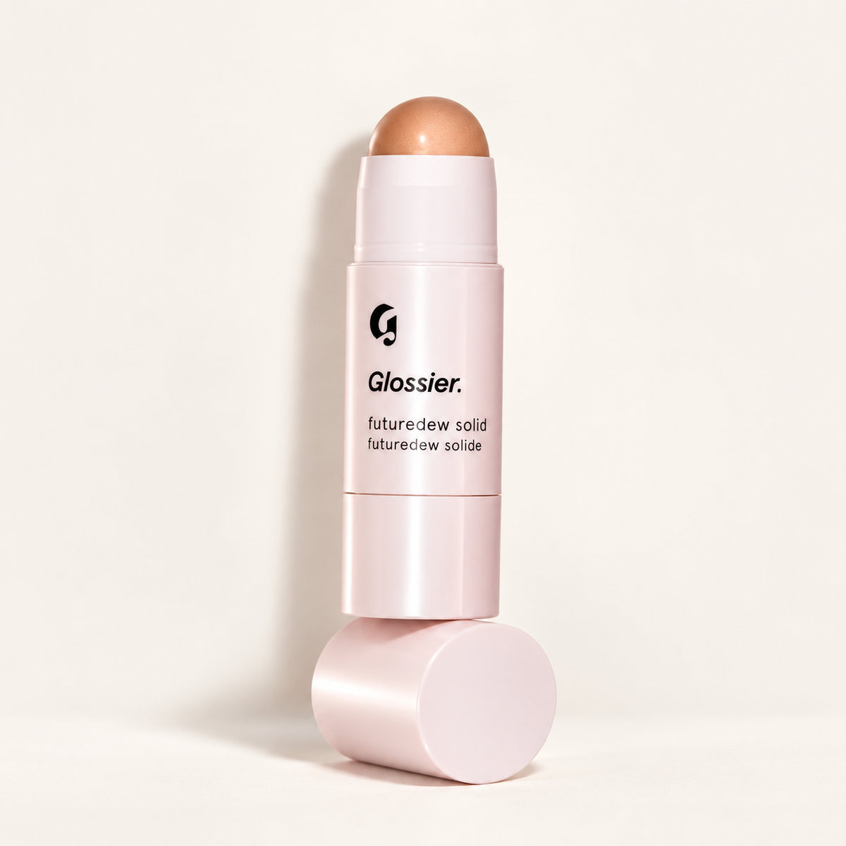 Glossier Futuredew Solid - sztyft ustawiony pionowo na różowej nakrętce, z kopułką w miedziano-brzoskwiniowym odcieniu, na jasnym tle | Ellemania