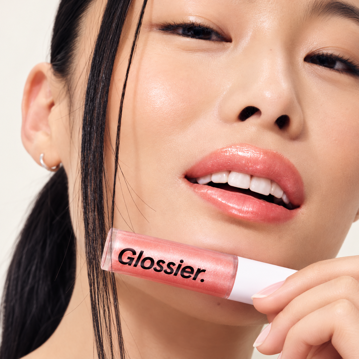 Glossier Lip Gloss Pink Rose - modelka z błyszczykiem na ustach trzyma przezroczystą tubkę z białą nakrętką przy brodzie | Ellemania