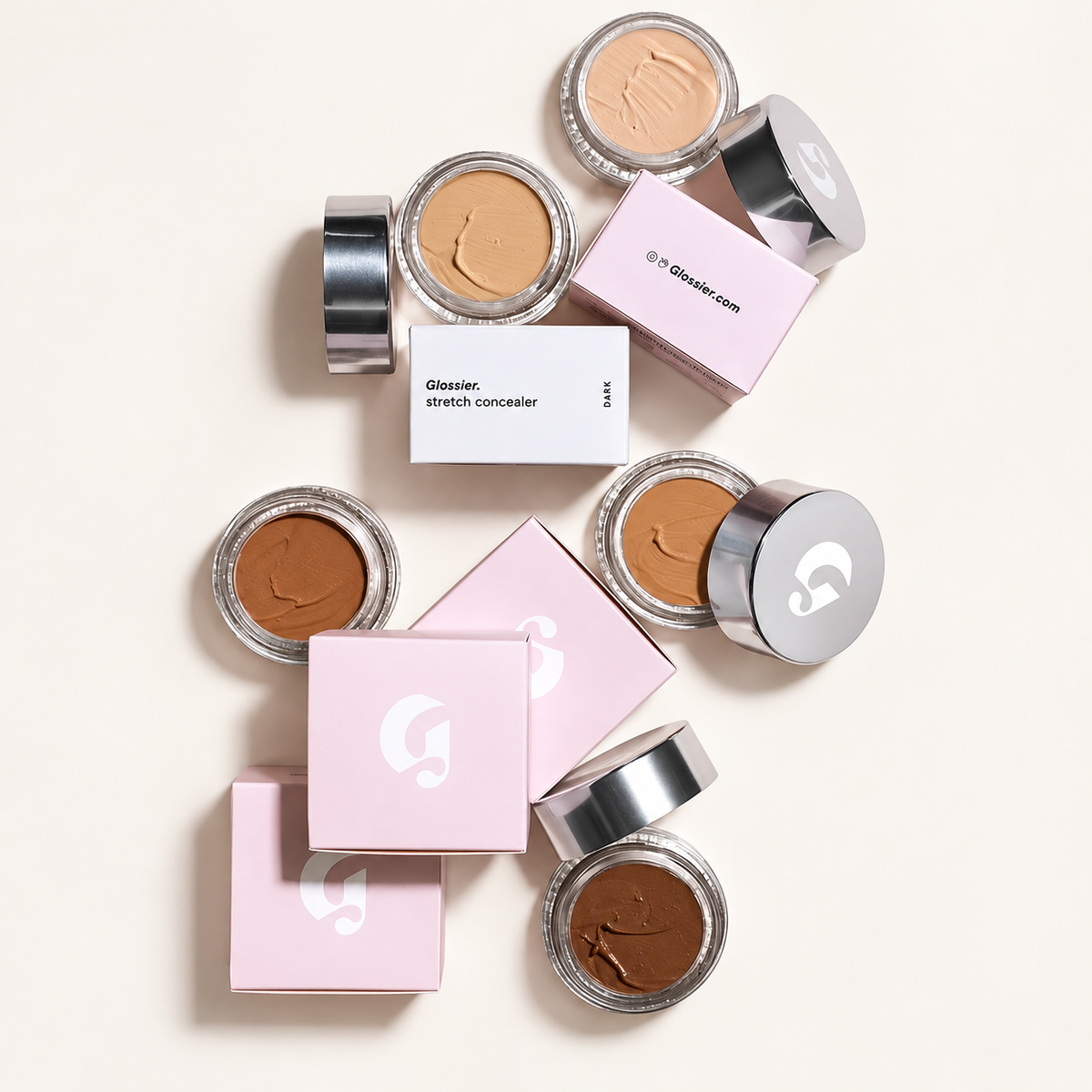 Glossier Stretch Balm Concealer - pięć otwartych słoiczków w odcieniach od jasnego beżu do ciemnego brązu, srebrne nakrętki z logo i różowe pudełka, na białym pudełku napis stretch concealer DARK, na jasnym tle | Ellemania