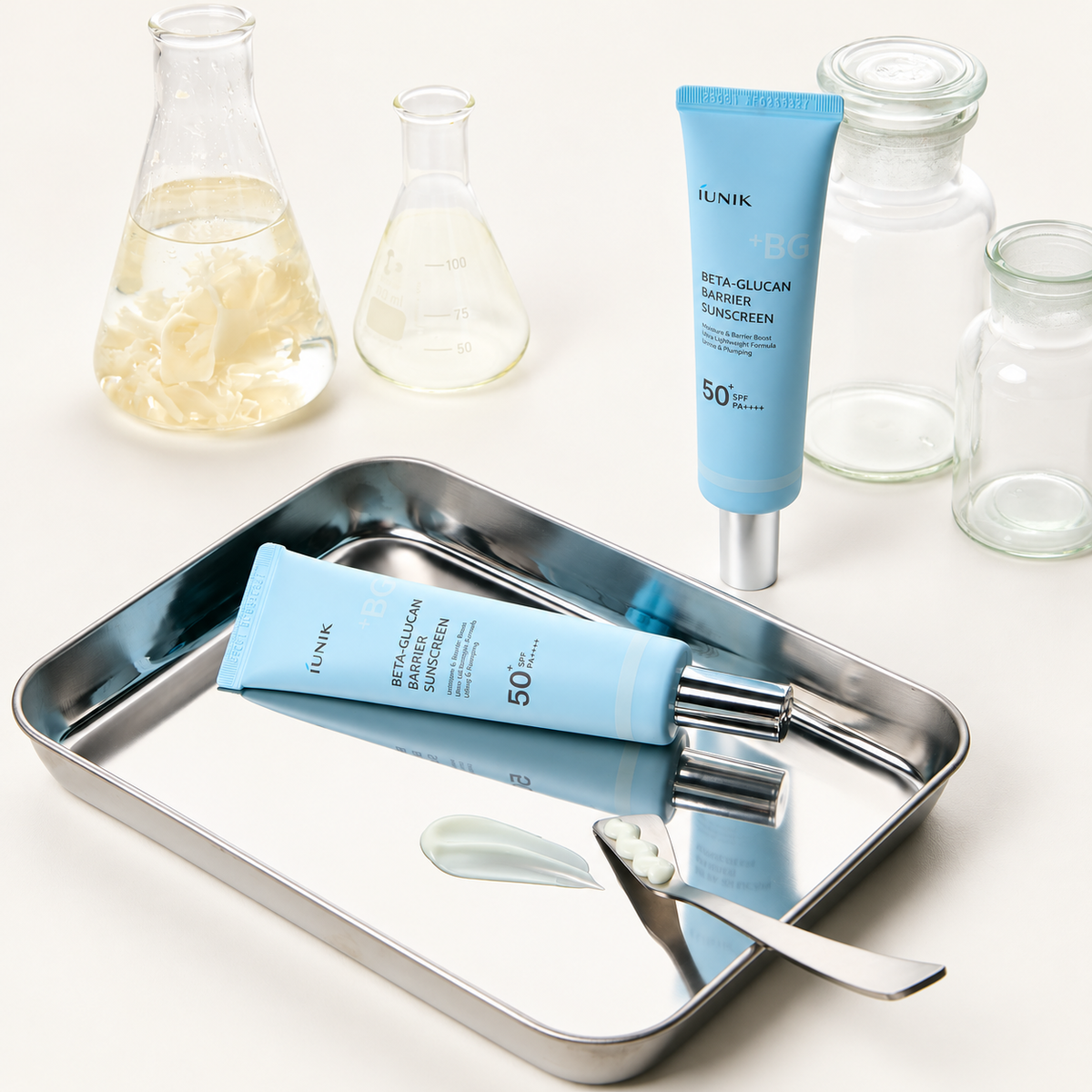 IUNIK Beta-Glucan Barrier Sunscreen - dwie tubki, jedna leżąca na metalowej tacy ze smugą kremu i szpatułką, w tle naczynia laboratoryjne | Ellemania