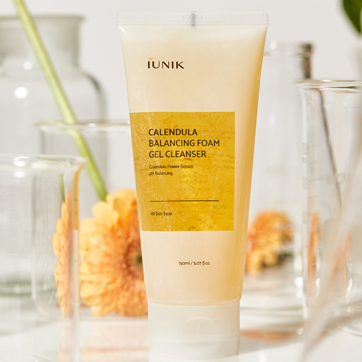 IUNIK Calendula Balancing Foam Gel Cleanser - tubka wśród szklanych naczyń laboratoryjnych i pomarańczowych kwiatów | Ellemania