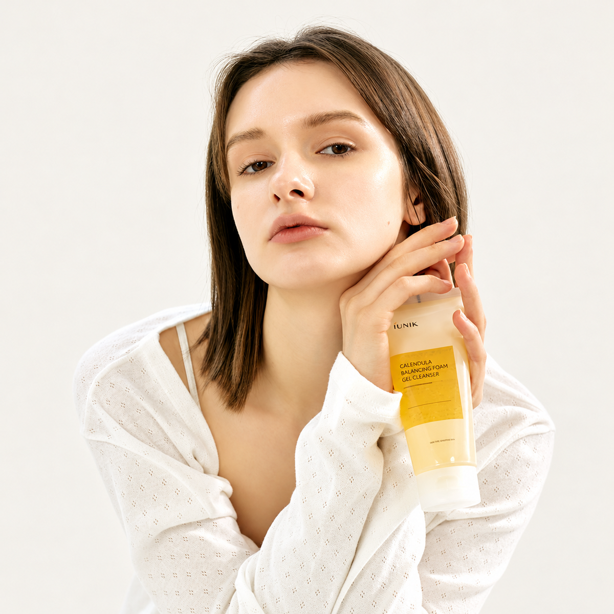 IUNIK Calendula Balancing Foam Gel Cleanser - modelka w białym ubraniu trzyma tubkę przy twarzy | Ellemania